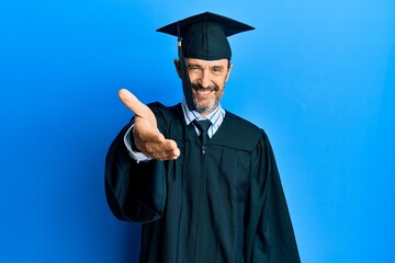 Middle age hispanic man wearing graduation cap and ceremony robe smiling friendly offering handshake as greeting and welcoming. successful business.