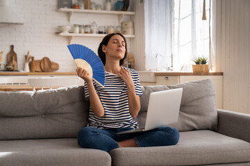 Tired millennial woman suffers from stuffiness and an inoperative air conditioner, waving blue fan...