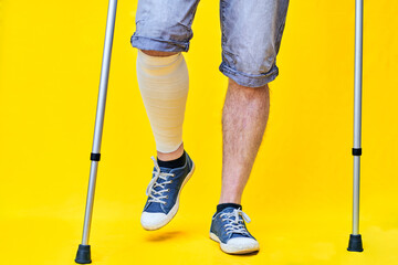 legs of a man from the front with shorts and on crutches, with a bandaged leg, on yellow background