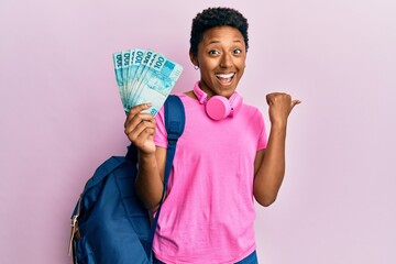 Young african american girl wearing student backpack and holding brazilian real banknotes pointing thumb up to the side smiling happy with open mouth