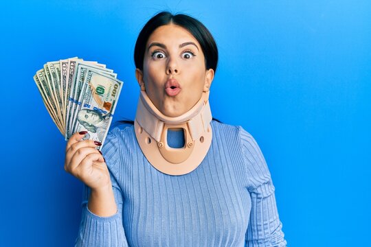 Beautiful brunette woman wearing cervical collar holding insurance money scared and amazed with open mouth for surprise, disbelief face