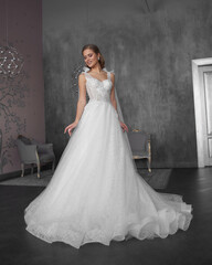 Full length bride portrait. Elegant female model wearing white fluffy wedding dress, standing and posing in grey room.