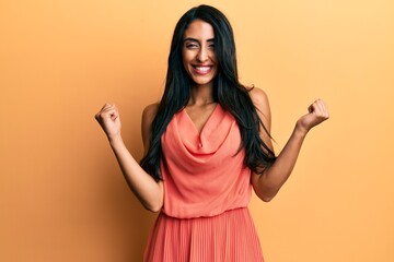 Beautiful hispanic woman wearing summer dress very happy and excited doing winner gesture with arms raised, smiling and screaming for success. celebration concept.