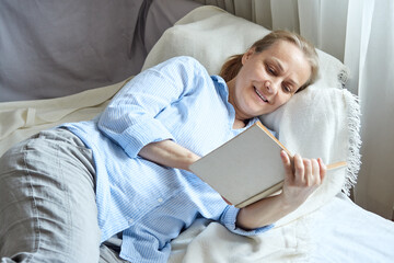 Smiling senior woman reading book while lying on sofa at home at free time.