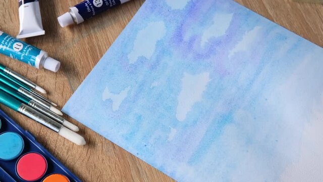 female artist's hand draws with colored paints, blue sky with clouds, background, abstract spots spread on white textured paper, watercolor painting, concept of creative art, art therapy