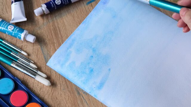 female artist's hand draws with colored paints, blue sky with clouds, background, abstract spots spread on white textured paper, watercolor painting, concept of creative art, art therapy