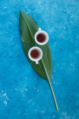 Two white cups with tea standing on a green leaf with blue background, view from top