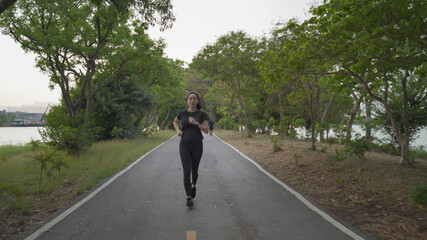 Obraz premium asian athlete woman in black sportswear and wireless headphone running in urban city trees park during sunset and bad cloudy weather. female jogging outdoor in evening. healthy workout.
