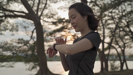 asian athlete woman using sport smart watch green nature park in evening sunset. female in sportswear touching screen application tracking activity and music. enjoy personal record data performance.