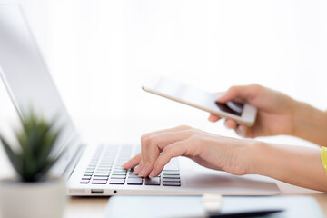 Closeup hand of young asian businesswoman working on laptop computer on desk at home office, freelance woman looking message on smart phone and typing on notebook, business and communication concept.