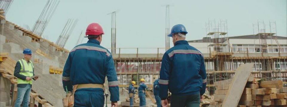 A Construction Site Workers with safety hard hats are walking through the commercial site. Camera following them from back.