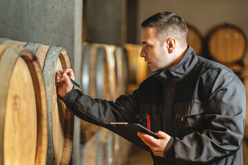 Side view of adult man winemaker at winery checking quality while standing between the barrels in...