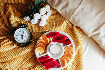 Various fruit for breakfast with clock showing 7 o'clock and rose flower on the bed