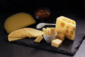 Several types of cheese in chunks, cut into slices and pieces in white bowl with jar of nuts on black stone board.