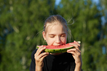Portrait of teenage girl with closed eyes in black T-shirt biting piece of watermelon, which holds with both hands in front of her on blurred natural background. Enjoy taste. Picnic in country