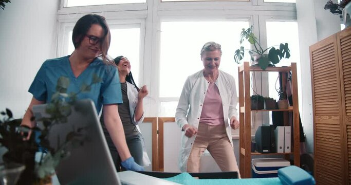 Three fun happy multiethnic female doctors dance together at work lab celebrating victory over coronavirus pandemic.