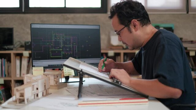 An Asian architect in a dark shirt is working on a design in his studio using a computer-aided design program and drafting board to drafting the construction plan.