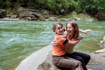 Fototapeta na wymiar Beautiful sisters have fun while sitting on the rock near mountains river. Two girl hugging and smiling on the big stone in forest in front of the big green river. Happy summer family vacation.
