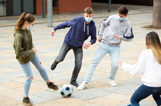 Positive teenagers in protective face masks are playing football in the school yard during a break in autumn day