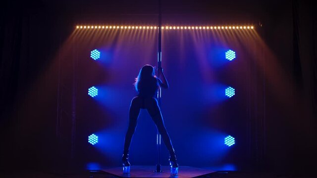 Rear view of sexy stripper pole dancing. Blonde erotically moves her hips, climbs a pole and spreads her legs in a split. Sensual pole dance in a dark studio with blue light. Silhouette. Slow motion.