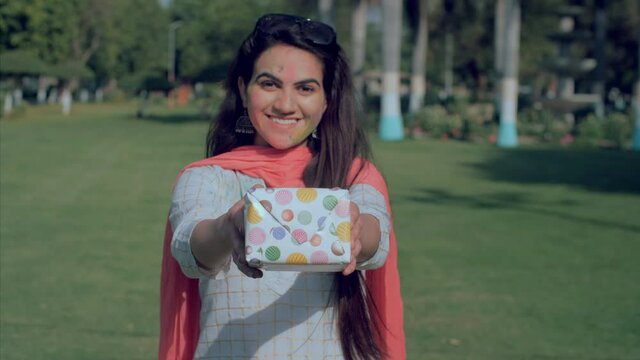 Lovely girl smeared with Gulal holding a colorful gift during Holi celebration. Medium shot of an Indian lady in white Kurti greeting people with presents on the occasion of the festival of colors