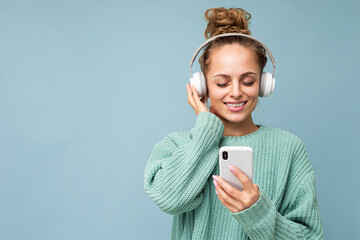 Photo of beautiful joyful smiling young woman wearing stylish casual clothes isolated over background wall holding and using mobile phone wearing white bluetooth headphones listening to music and