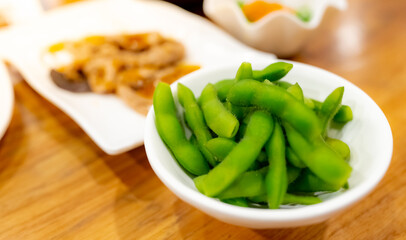 Edamame or green soybeans blanched in white bowl on wooden table at Japanese restaurant. Pods of green soy beans on blurred Japanese food. High fiber and protein food. Plant-based protein. Vegan diet.