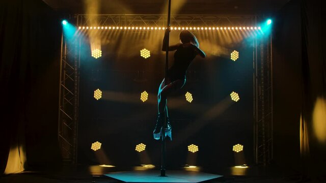 Attractive striptease dancer performs tricks on a pole. Silhouette of a sexy blonde woman in lingerie and high heels. Exotic dance in a dark studio with smoke and neon lighting. Slow motion.