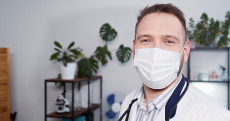 Healthcare expert portrait. Happy doctor man in white coat and face mask smiles at camera at modern hospital office.