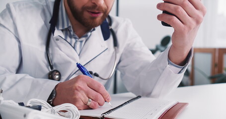 Close-up young Caucasian male doctor consulting remote patient using smart phone web conference, taking notes at office.