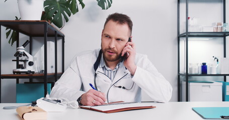 Serious young male Caucasian doctor consulting remote patient on self isolation talking on the phone at modern office.