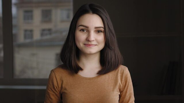 Portrait of lonely happy cute teenager girl standing indoors posing in room looking at camera. Close-up caucasian millennial friendly brunette woman smiling with calm carefree female face expression