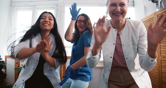 End of fighting coronavirus. Zoom out three fun multiethnic happy female doctors dancing together at clinic lab office.