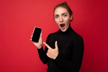 Photo of surprised attractive positive young brunette woman wearing black sweater standing isolated over red background showing mobile phone with empty screen for mockup looking at camera pointing
