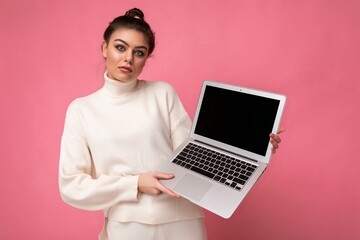 Photo of beautiful prette brunette young woman with gathered dark hair wearing white sweater holding computer laptop and looking at camera isolated on pink wall background