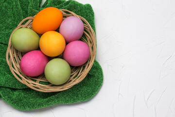 The Easter eggs in the basket flat lay on a grass-green tablecloth on a white textured background with a copy of the space
