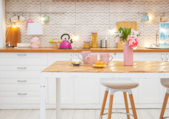 Pink vase with flowers and mugs for tea on the table in a light kitchen