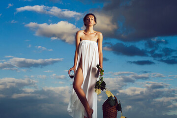 Young woman in white sexy dress with red wine in glass wicker bottle vine and grapes on nature over blue sky. Wine Glasses And Bottle With Barrel In Vineyard. Winery concept.