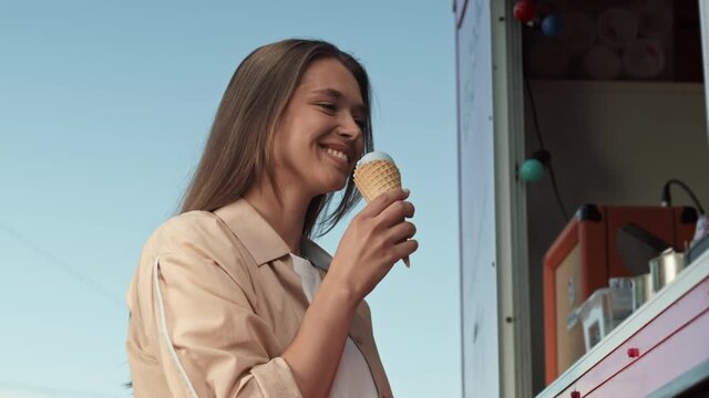 Low angle chest-up POV of young Caucasian woman standing by food truck outdoors, buying ice cream in waffle cone, then tasting it, looking and smiling on camera