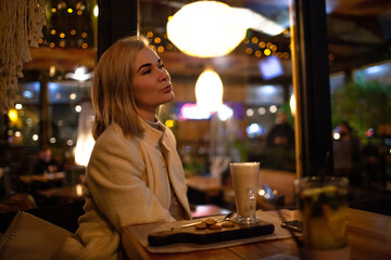 Beautiful woman sit in night cafe and eat fast cafe and dink coffee in evening.