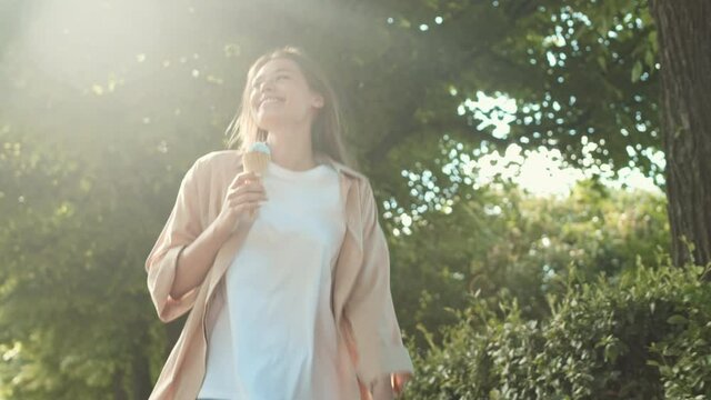 Medium tracking POV of happy playful Caucasian woman, walking jumping in summer park, holding ice cream in cone, smiling and looking on camera