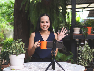 Asian  woman coach or speaker, sitting at table in garden, record live video broadcast on modern...