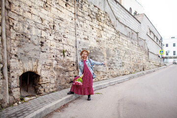 Fashion style woman dressed jeans jacket, hat and red dress walking in the street of old city with bouquet of flowers