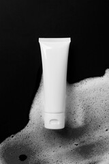 White plastic tube mockup with moisturizer cream, shampoo or facial cleanser on black background with soft soap foam, top view. Treatment spa beauty skincare cosmetic product
