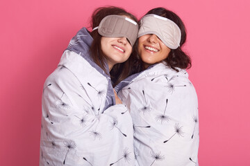 Horizontal shot of two young girls, poses with sleeping mask over eyes, wrapped white blanket against rosy wall, two friends with smiles hugging on pajama party, being ready to fall asleep.