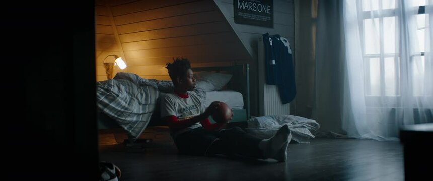 WIDE Bored African American Black kid teenager boy playing with football ball in his attic bedroom at home. Shot with 2x anamorphic lens