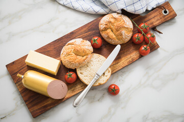 Feine Kalbsleberwurst mit zwei Weizen Rosen Brötchen, Tomaten, Butter und Messer auf Holzbrett, Geschirrtuch und Marmor Hintergrund als Brotzeit Vesper