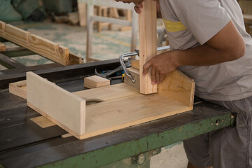 A young male carpenter builder in a gray T-shirt and working overall equals a wooden bar with a milling machine in the workshop, in the background wooden boards, in the workshop