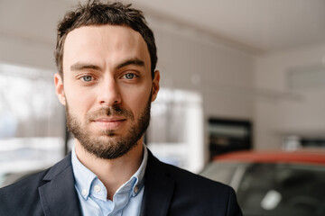 Unshaven white man looking at camera while standing in car showroom