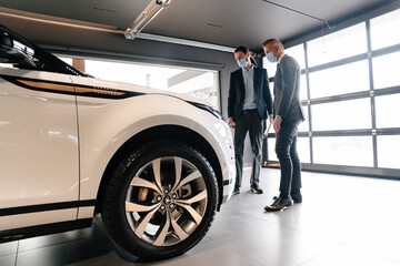 Mid white manager in face mask showing car to customer in showroom